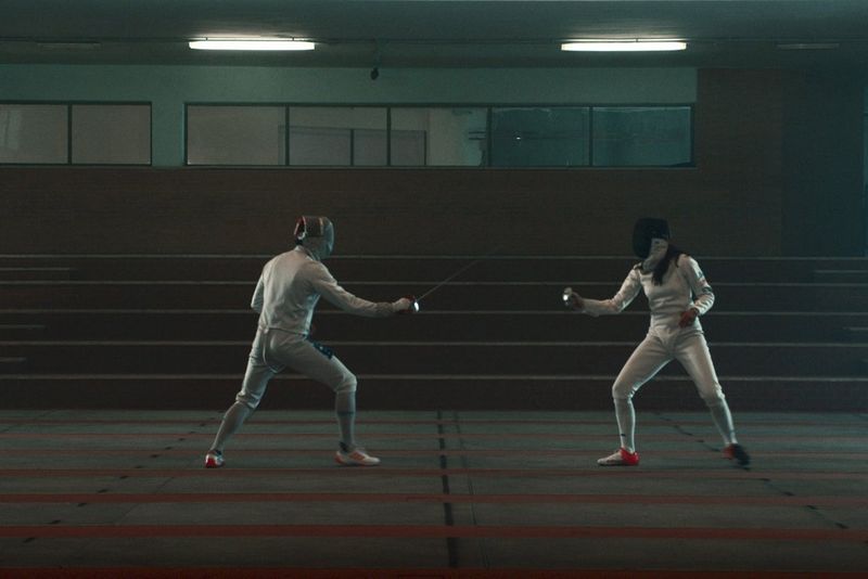 Two fencers sparring in a room lit dimly with fluorescent lighting, one wearing a black mask, the other a silver one.