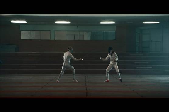 Two fencers sparring in a room lit dimly with fluorescent lighting, one wearing a black mask, the other a silver one.