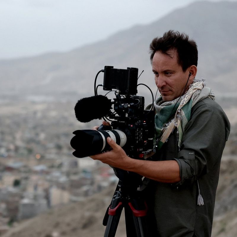 A man standing on a hillside with a Canon camera on a tripod. A sprawling city stretches into the distance behind him.