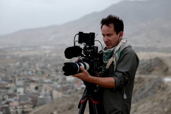 A man standing on a hillside with a Canon camera on a tripod. A sprawling city stretches into the distance behind him.