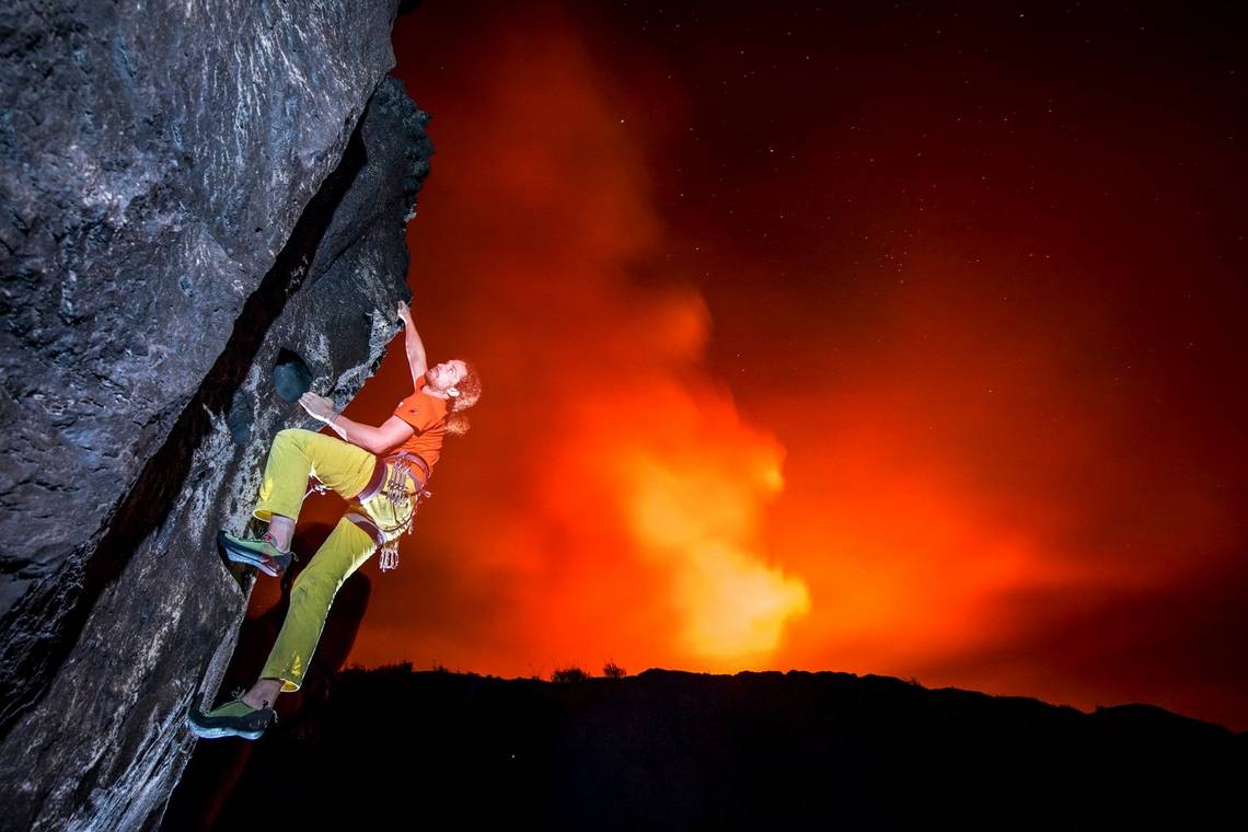 A rock climber scaling the side of an active volcano, with the red glow of the caldera behind. 