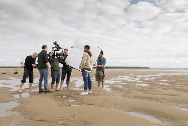 A group on a beach seen from a distance, with one filming with a camera in a gimbal while two others hold a large reflector.