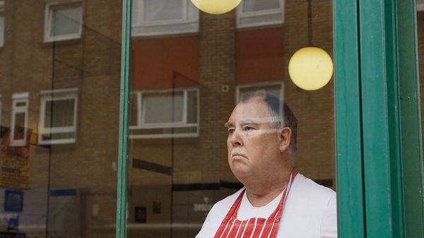 The owner of a pie and mash shop, wearing a red apron, looks out of his shop window. A film still from Zed Nelson's The Street.