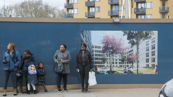 People standing on the pavement in front of a billboard advertising new luxury apartments that are being built. A film still from Zed Nelson's The Street.