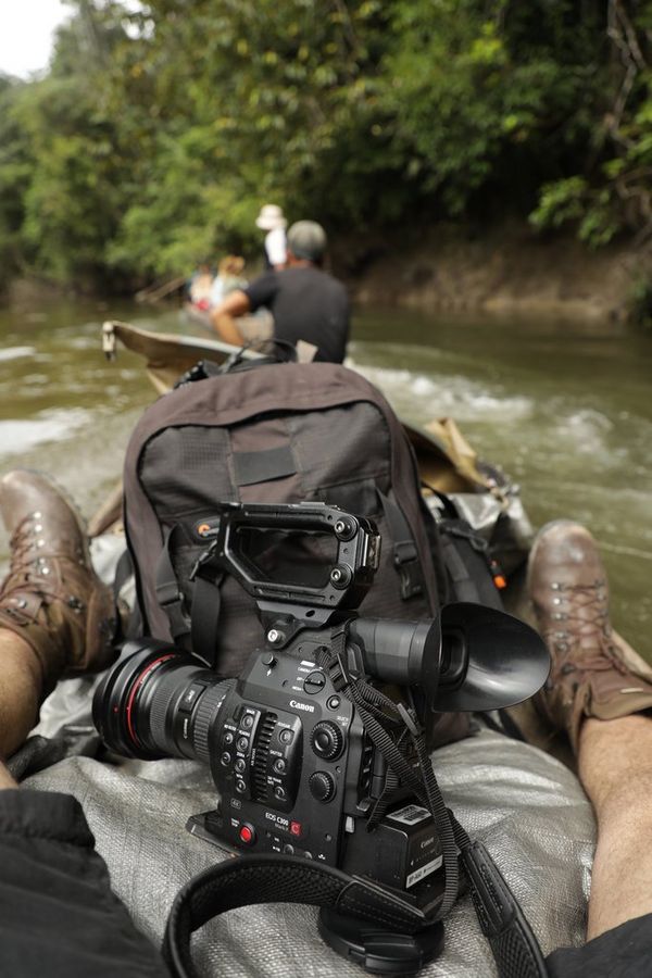 A Canon EOS C300 Mark II sits between a man's legs in a canoe on a river through dense jungle.