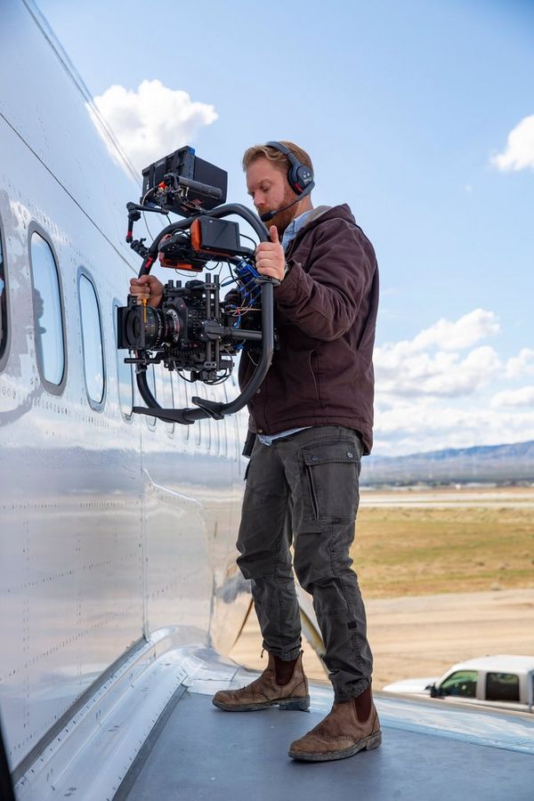 Cinematographer Steve Holleran stands on the wing of an aircraft holding a Canon EOS C300 Mark III on a gimbal and films through the window.