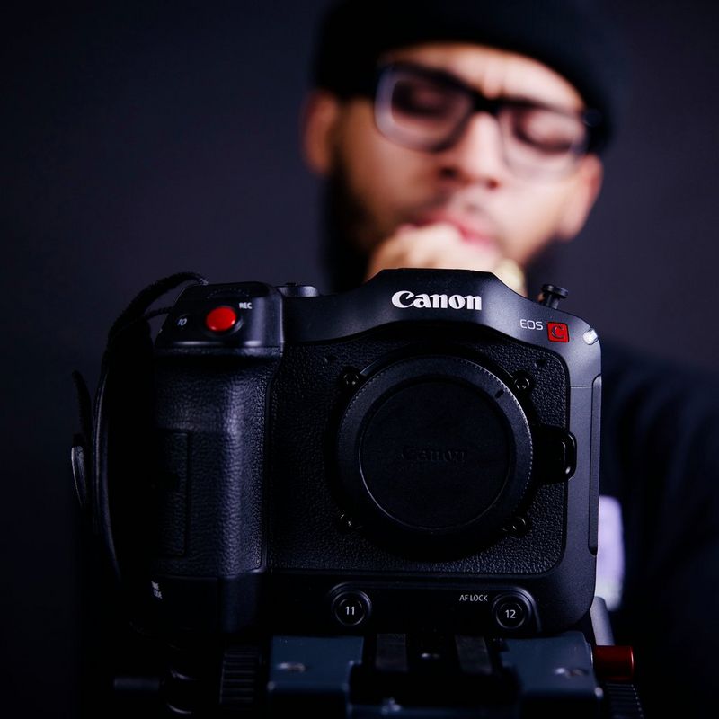 A close-up of a Canon EOS C70 with a bespectacled figure standing behind it.