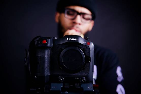 A close-up of a Canon EOS C70 with a bespectacled figure standing behind it.