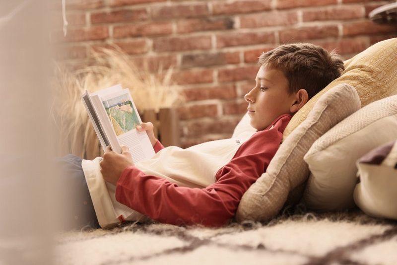 A young boy reads a book while leant against a large stack of cushions.