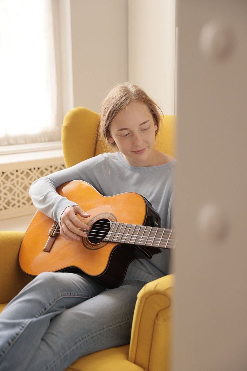 Through a doorway, a young woman can be seen sitting in a yellow armchair playing the guitar.