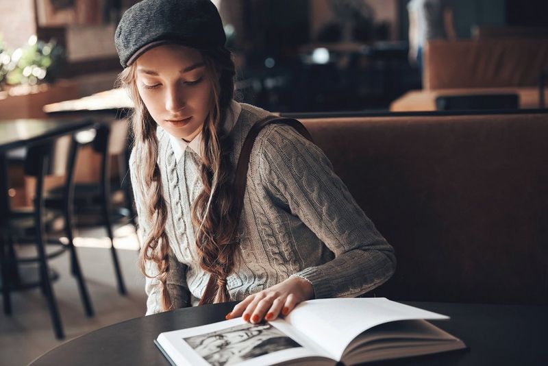 A woman wearing a jumper and a cap sits in a cafe reading a book.