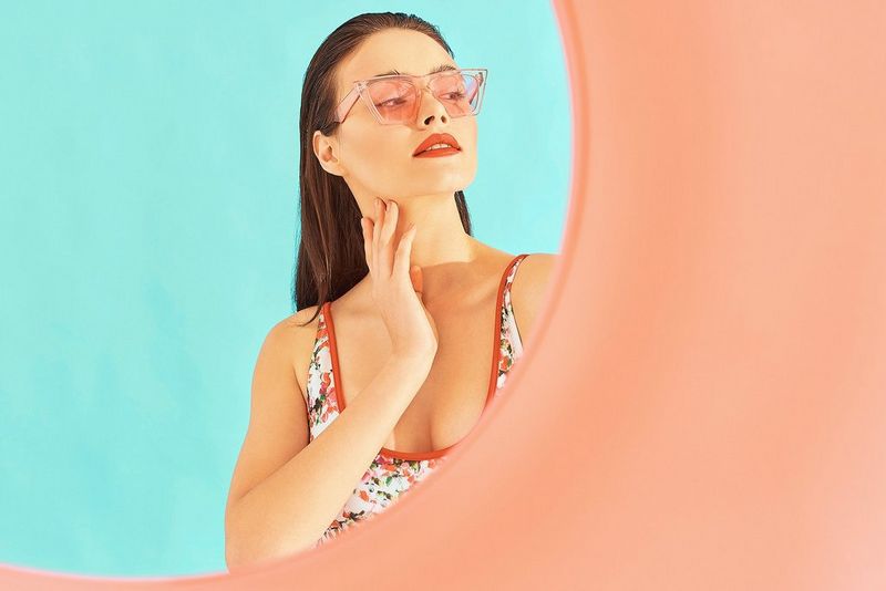 A woman in pink sunglasses stands in front of a blue wall, seen through a pink tunnel.