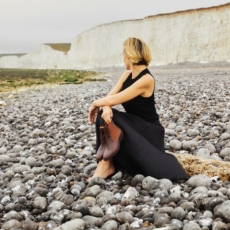 A woman looks away from the camera as she sits at a pebbly beach while holding a pair of boots, taken on a Canon EOS 5D Mark IV by Kid Circus. 