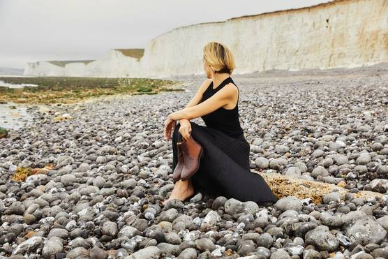  A woman looks away from the camera as she sits at a pebbly beach while holding a pair of boots, taken on a Canon EOS 5D Mark IV by Kid Circus.