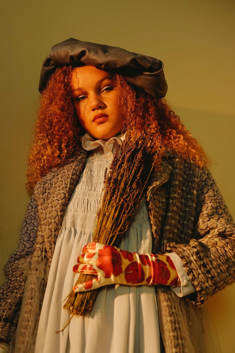 A portrait of a woman with curly, ginger hair holding a bunch of dried lavender in her gloved hand. 