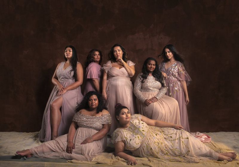 A group shot of plus-size Asian models Michelle Elman, Bishamber Das, Kat Henry, Mina Kumari, Simsimma Sandhu, Saalene Sivaprasad and Vanessa Sison wearing long, floaty dresses in pastel shades. 
