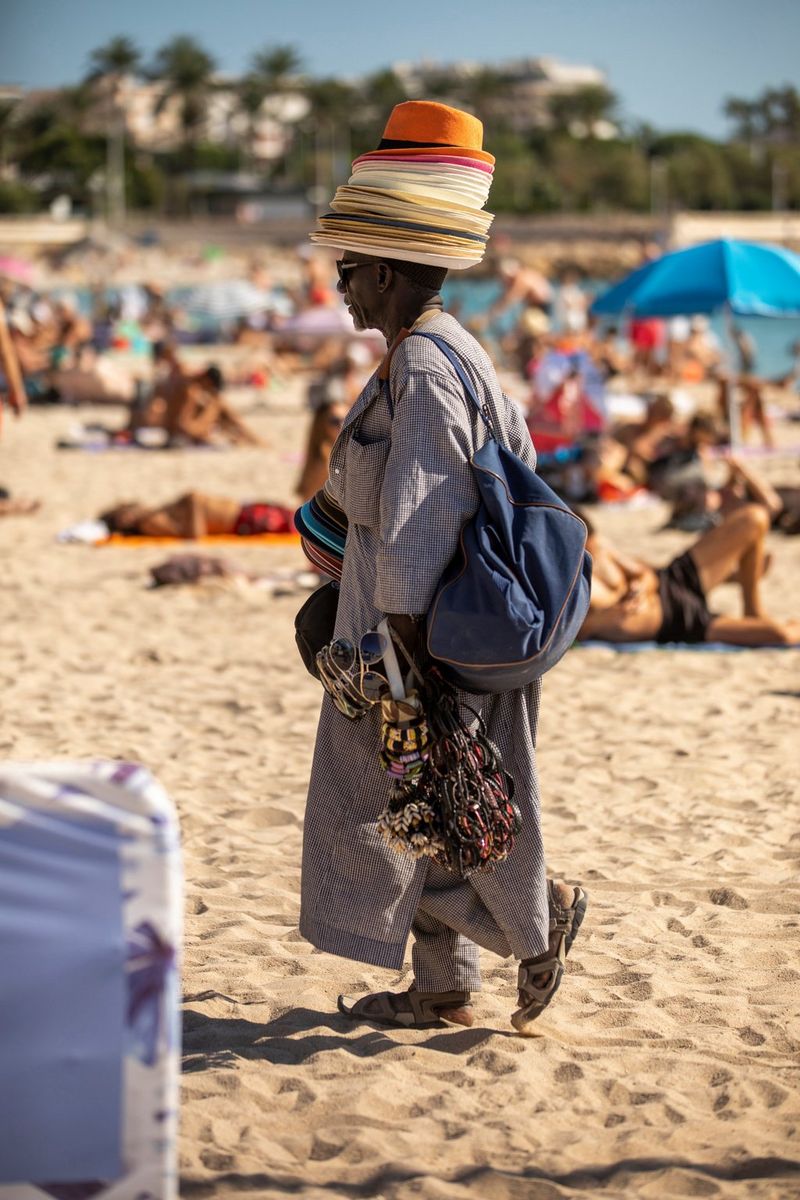 A beach seller walks across the sand carrying bags and accessories and wearing a pile of straw hats.