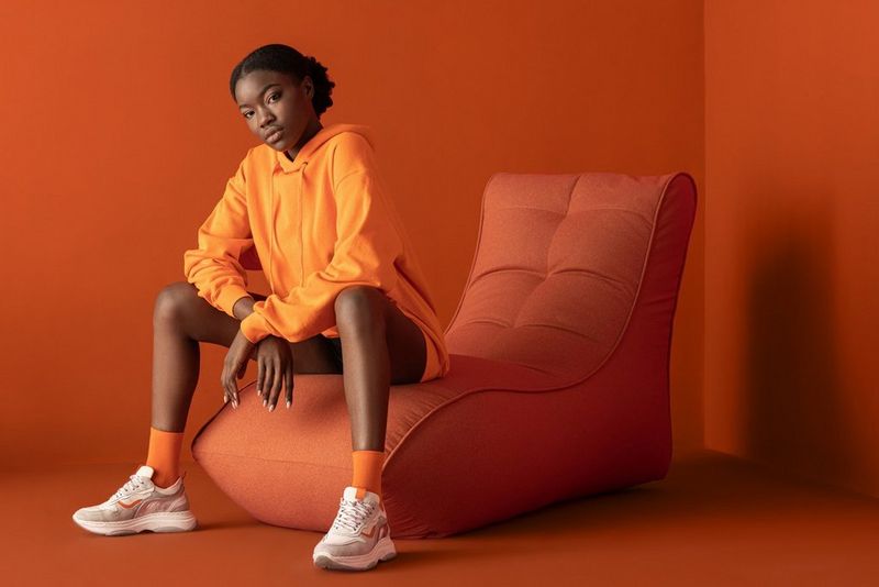 A woman wearing an orange hoodie and orange socks sits on an orange chair against orange walls and floor, leaning forward to rest her elbows on her knees and looking at the camera. Taken by Jaroslav Monchak on a Canon EOS R5.