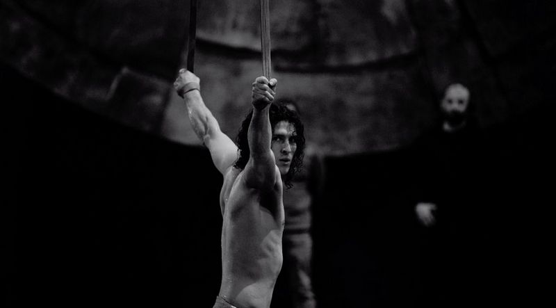 A black-and-white still from the Cirque du Soleil film of a bare-chested male acrobat hanging from aerial silks.