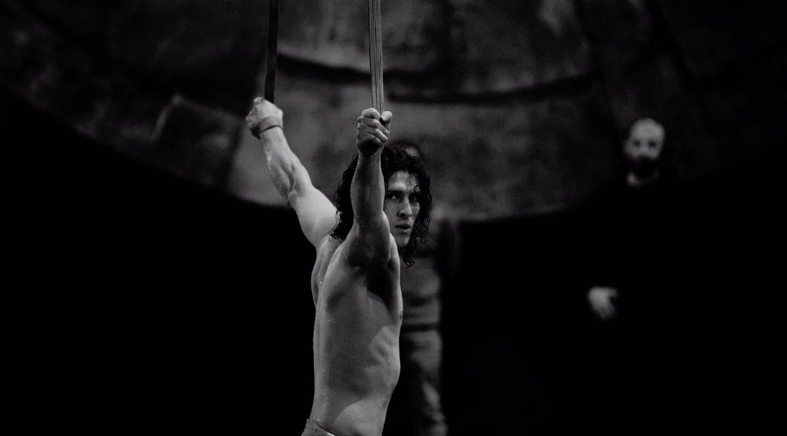 A black-and-white still from the Cirque du Soleil film of a bare-chested male acrobat hanging from aerial silks.