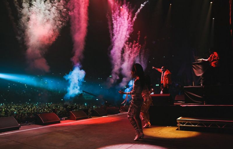 Dancers on the stage at a music concert, with multicoloured trails of smoke rising above the crowd.