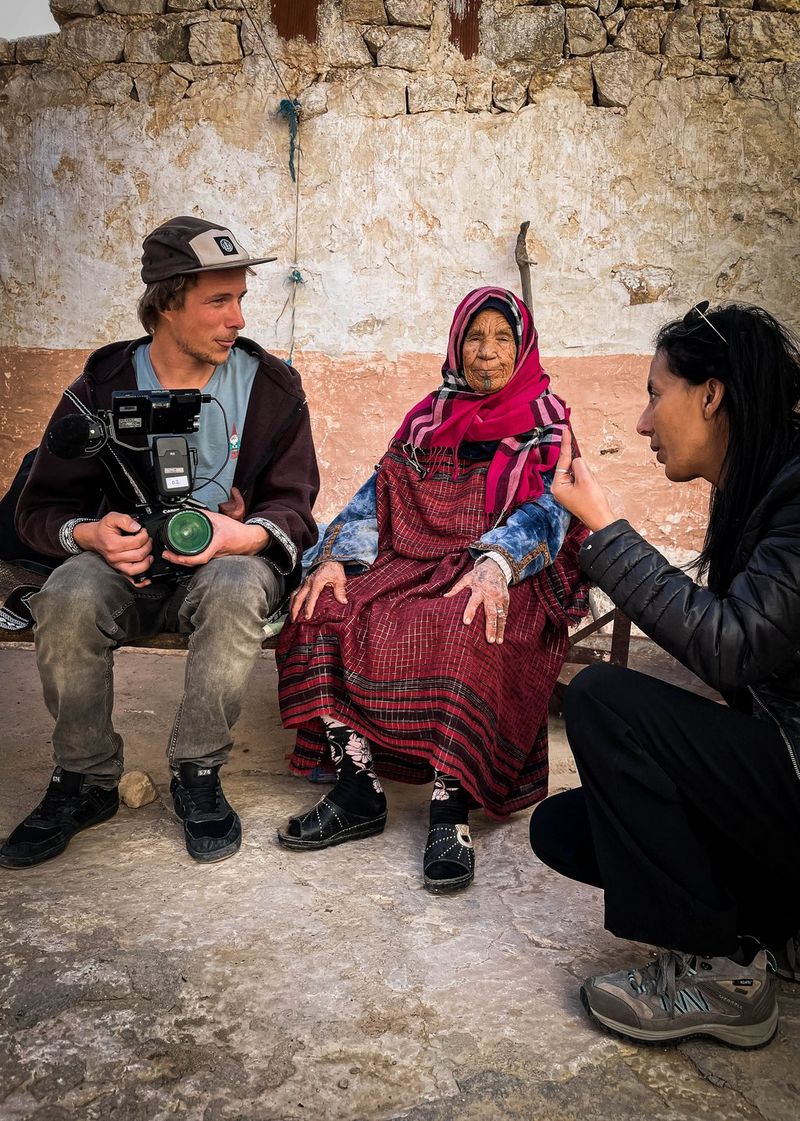 Filmmaker Michael Zomer holds a Canon camera and sits and chats with two people one of whom is dressed in clothing indigenous to the area. 