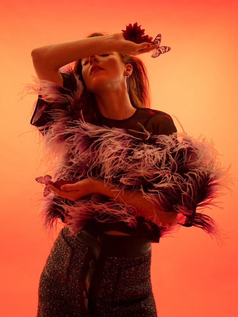 A woman in a feathered top poses in front of an orange and yellow backdrop, her arm raised to obscure her face.