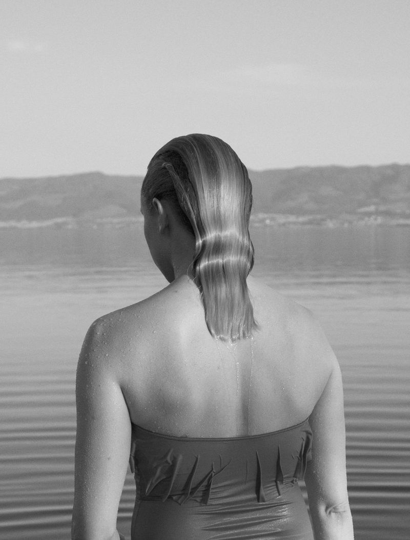 A woman looking at an expanse of water with her back to the camera. She has wet hair and is wearing a swimsuit.