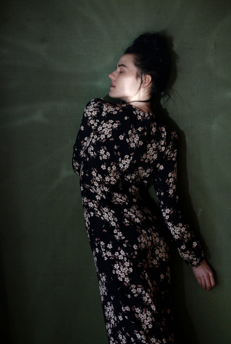 A side profile of a woman in a floral patterned dress leaning against a green wall.
