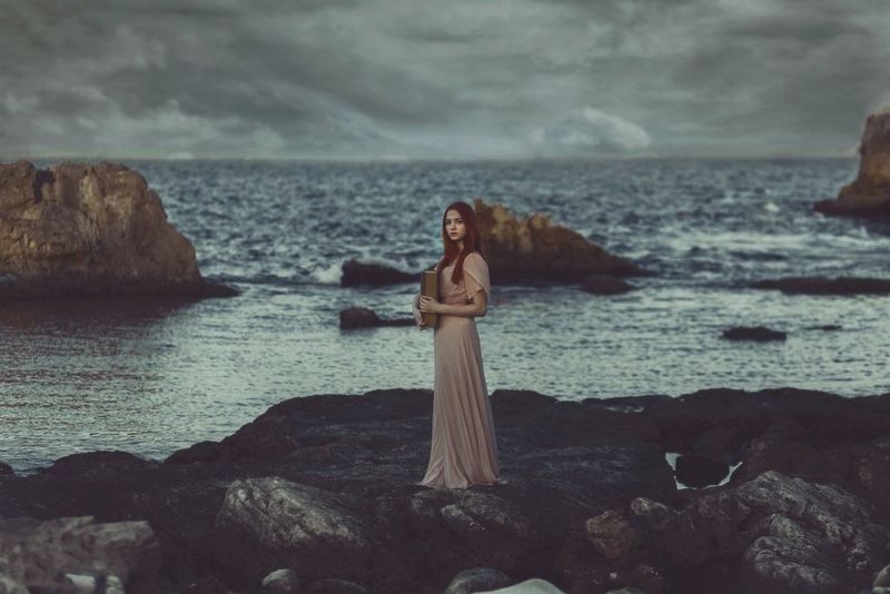 A model in a long pink dress holds a book on a rocky beach. Waves lap at the shore behind her.