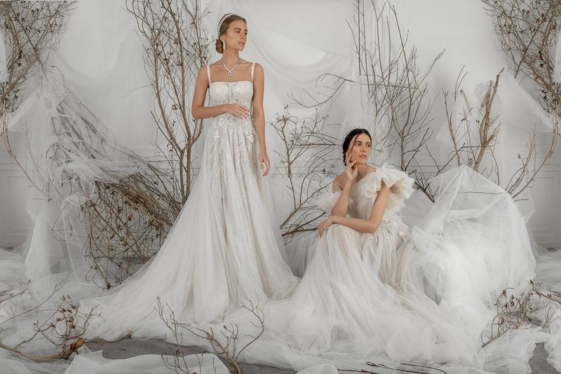 Two women wearing flowing white gowns surrounded by white netting and tree branches. 
