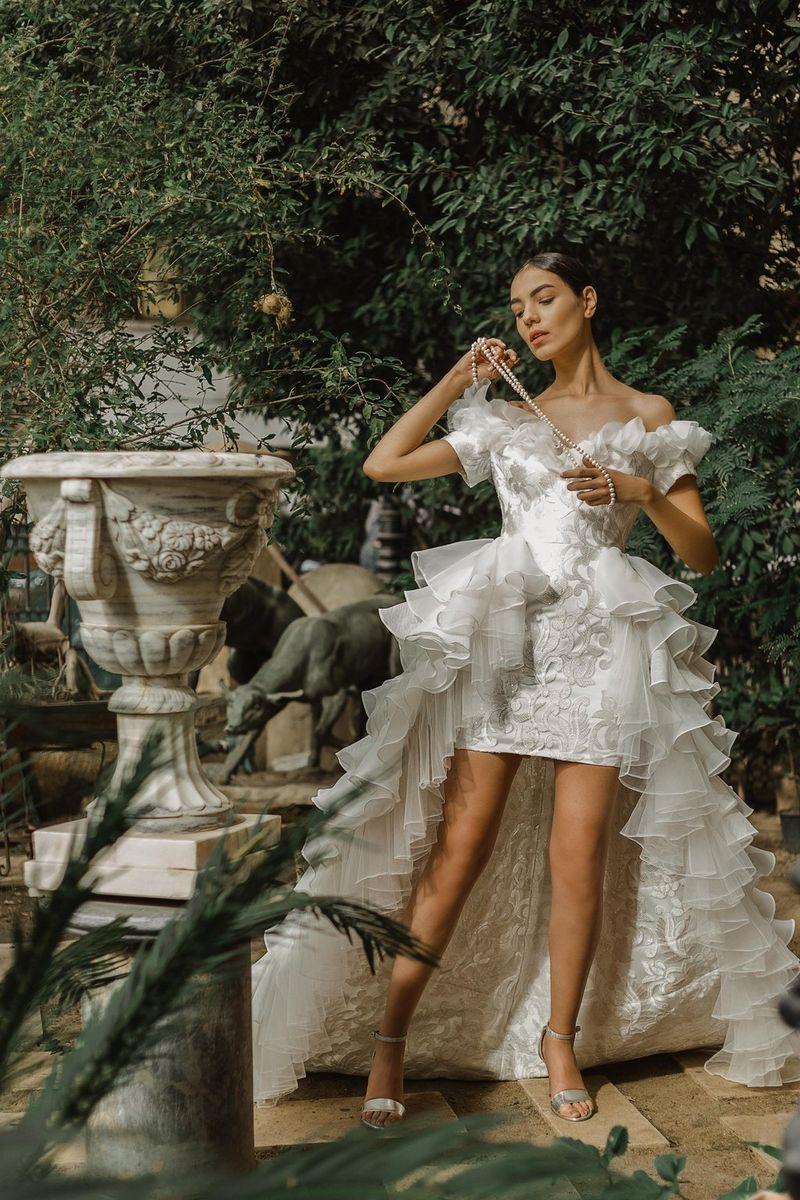 A model in a short white dress with a long ruffled train poses in a garden next to a large urn.