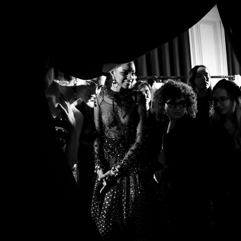 A black-and-white shot of models watching a fashion show from behind a curtain in their dressing room. 