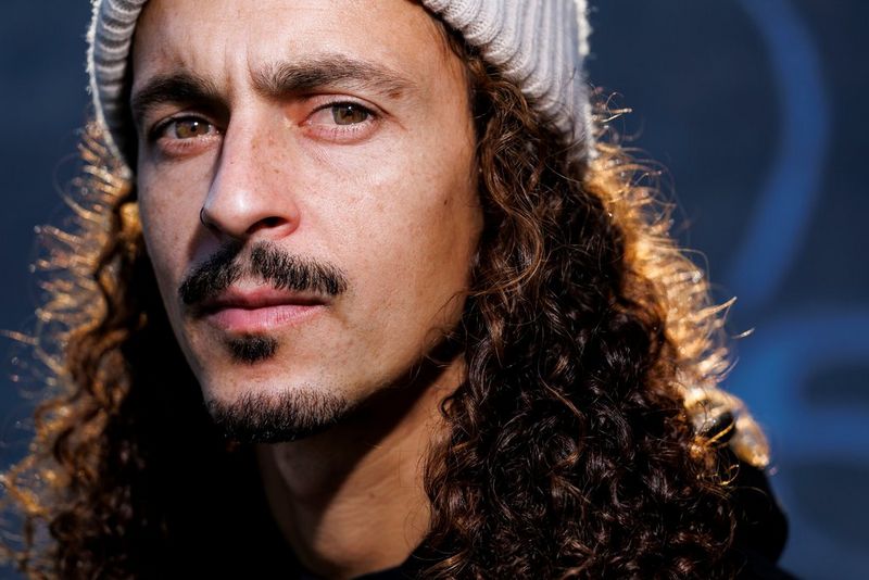 A close-up portrait of a man with short facial hair, long curly hair and a woolly hat.