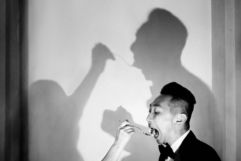 Scatto in bianco e nero di un uomo in giacca che mangia una cucchiaiata di torta. Le ombre sulla parete alle sue spalle completano la scena, rivelando che il cucchiaio è tenuto da qualcun altro.