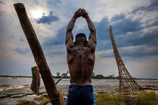 A muscular young man with his arms raised aloft and hands linked stands on a riverbank. Taken by Brent Stirton on a Canon EOS R5.