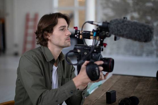 Filmmaker and activist Jack Harries leans on a wooden table setting up a shot with his Canon camera rigged with a mic.
