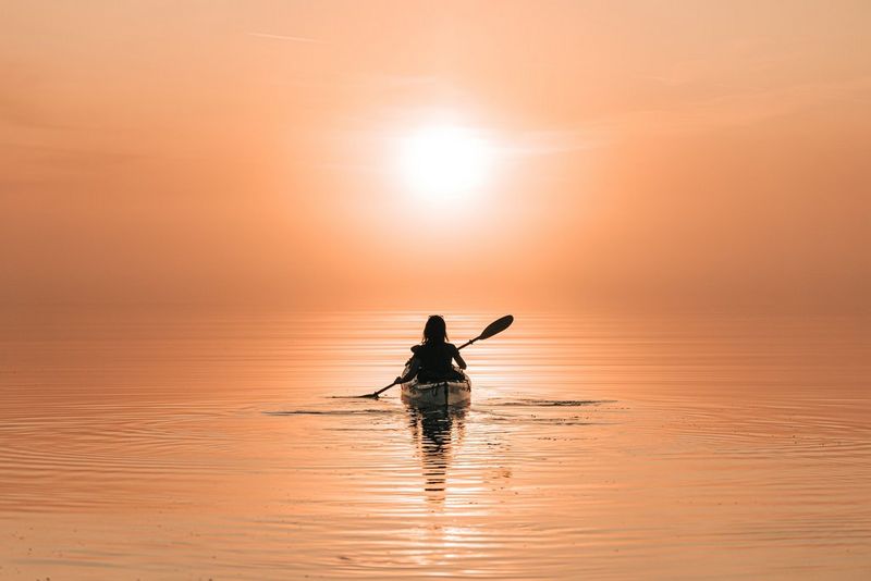 A figure in a kayak paddles out towards the horizon, the water and sky bathed in an orange hue.