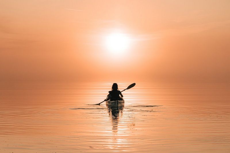 A person kayaking on still water, in silhouette, with the setting sun turning the water orange. 