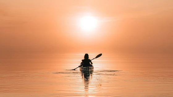 A person kayaking on still water, in silhouette, with the setting sun turning the water orange. 