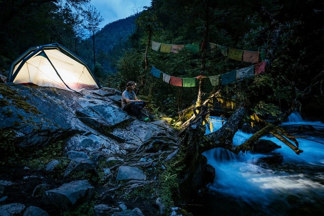 Мужчина сидит возле своей палатки, установленной на камнях у водопада.