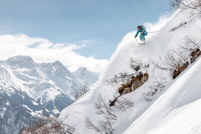 Лыжник, одетый в бирюзовый костюм, на краю крутого спуска на фоне заснеженных гор.