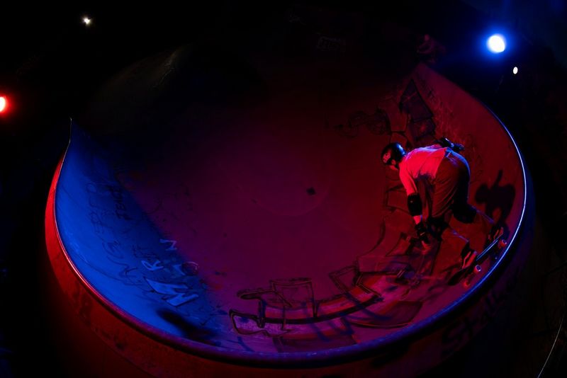 Созданная с помощью объектива «рыбий глаз» фотография девочки, катающейся на рампе в скейт-парке в ночное время среди источников красного и фиолетового света.