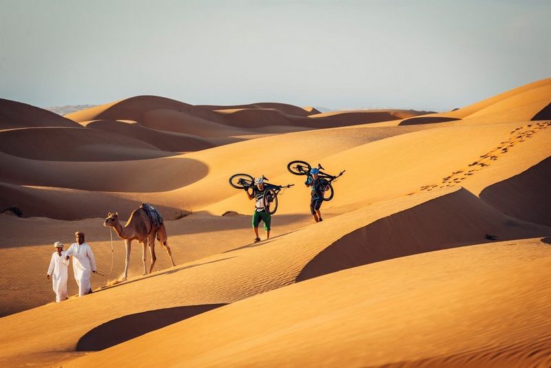 Двое мужчин несут на плечах велосипеды, следуя по пустыне за двумя мужчинами в арабской одежде и верблюдом.