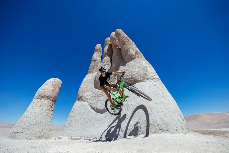 Мужчина на горном велосипеде выполняет трюк на огромной каменной статуе вскинутой вверх руки.