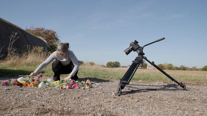 Cinematographer Tania Freimuth crouches down to arrange colourful flowers on a gravel road. A Canon EOS C70 camera is set up on a tripod to film the flowers.