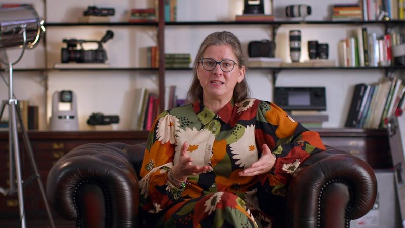 Filmmaker Tania Freimuth sits in a leather armchair in front of shelves filled with cameras, lenses and books.