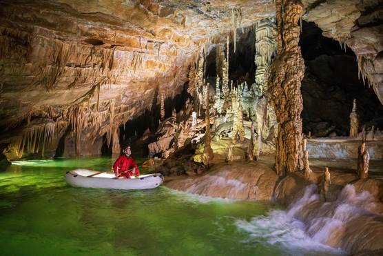 A man sits in a small inflatable boat inside a stunning cave lake, with bright green water and overhanging rocks.