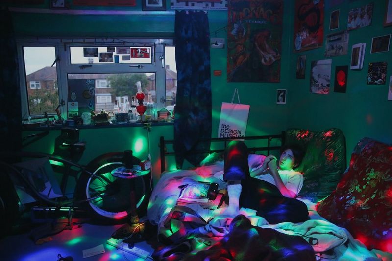 Jeune homme couché sur un lit, dans une chambre en désordre, éclairée par des lumières multicolores émanant d'un projecteur.