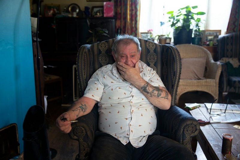 Homme assis sur un fauteuil, une main sur sa bouche, l'autre tenant une pipe, avec des tatouages visibles sur les bras.